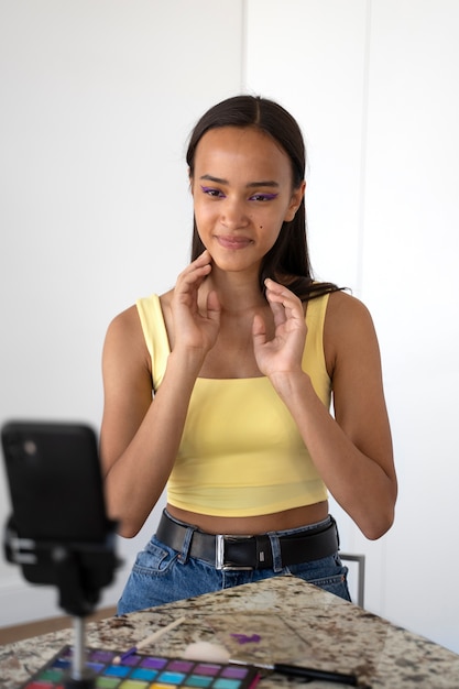 A close-up shot of a young Brazilian digital influencer filming a makeup tutorial in a well-lit studio. The influencer is smiling and engaging, demonstrating various makeup products and techniques. The background includes professional lighting equipment and a clean, organized workspace.